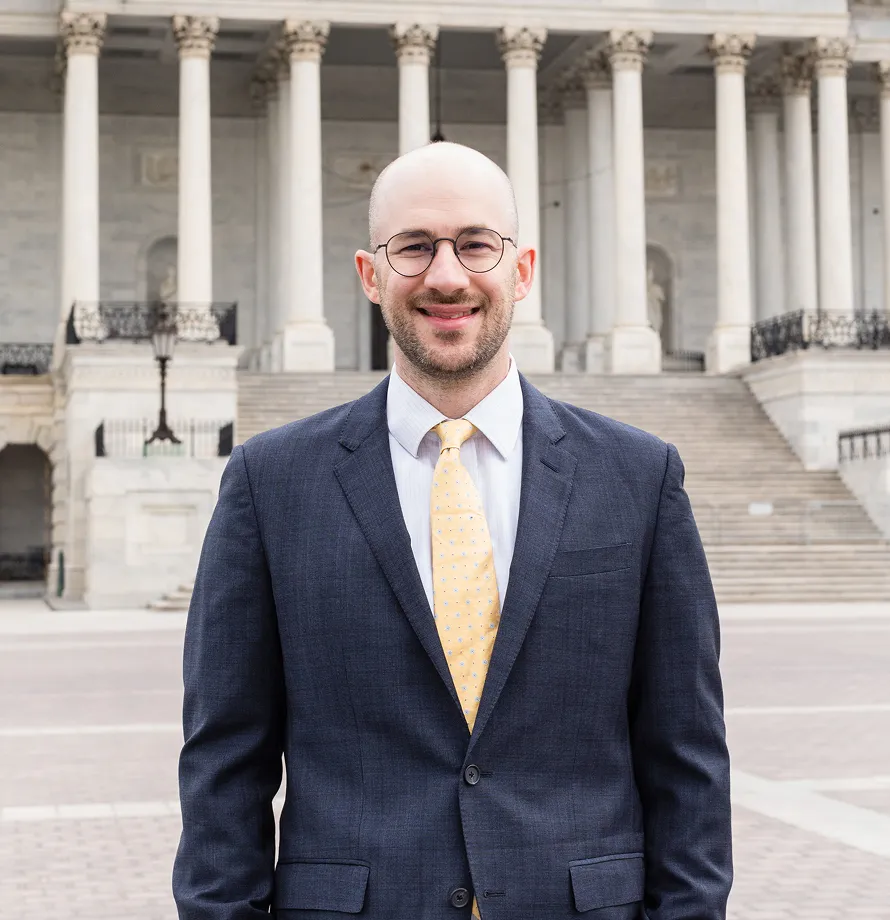 Professional man wearing glasses, navy suit, and yellow tie standing in front of neoclassical building with columns.