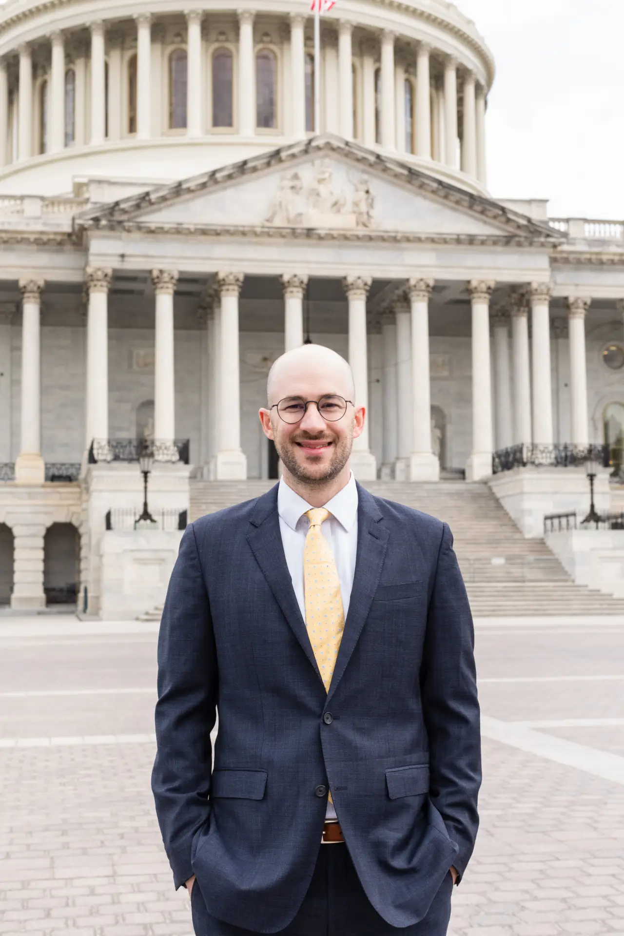 Professional man in dark suit and yellow tie standing in front of a classical government building with columns.