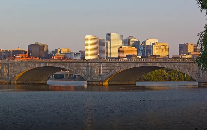 Stone arch bridge over calm river with modern city skyline and evening light.