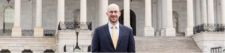 Man in dark suit and yellow tie standing in front of a large stone building with columns and steps.