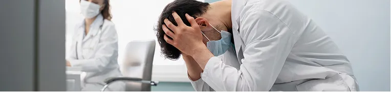 Two medical professionals in white coats and masks, one seated with head in hands.