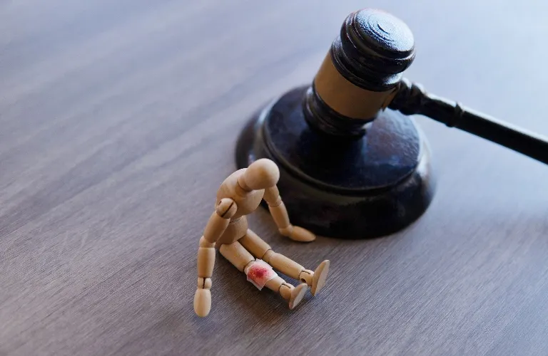 Wooden mannequin with red injury mark on knee sitting beside black judge's gavel on wooden surface.