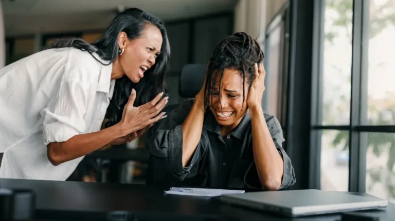 Two women in an office; one angrily yelling, the other covering ears in distress.