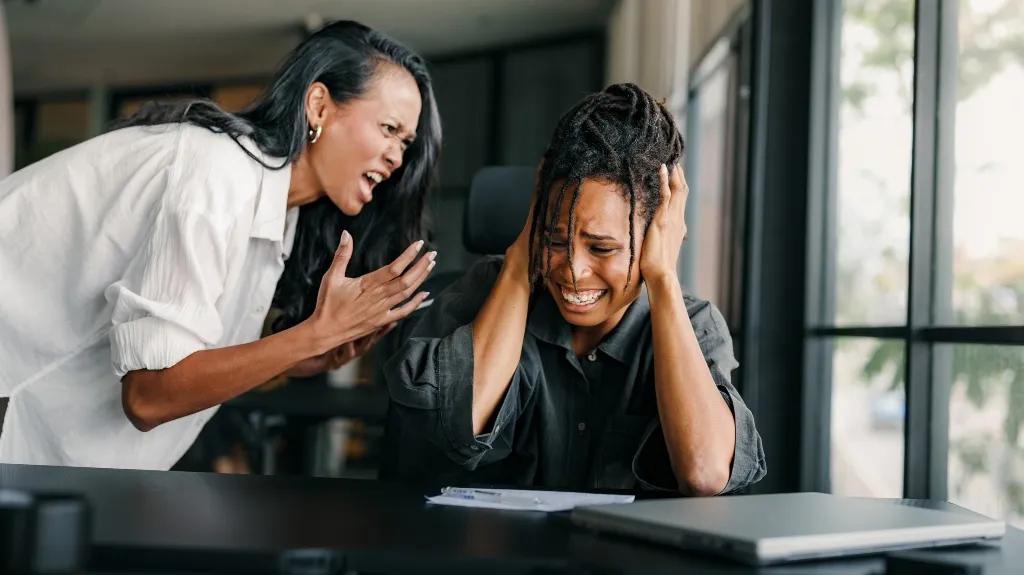 Two women in an office; one angrily yelling, the other covering ears in distress.