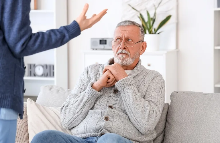 Elderly man in gray sweater sitting on couch looking concerned at standing person gesturing.
