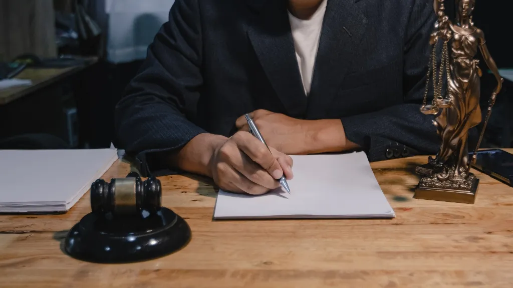 Person in dark suit writing on paper at wooden desk with gavel and Lady Justice statue.