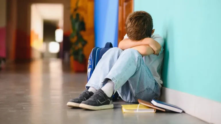 Young boy sitting on the floor with head buried in arms, blue backpack and books beside him.