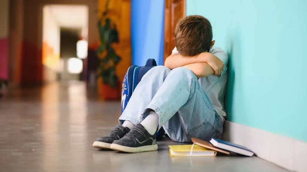 Young boy sitting on the floor with head buried in arms, blue backpack and books beside him.