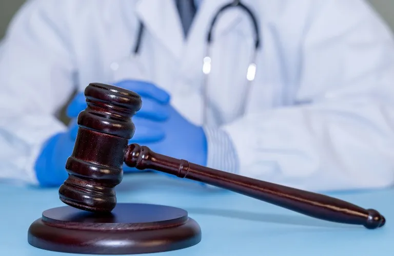 Wooden judge's gavel in foreground with person in medical coat and blue gloves in background.