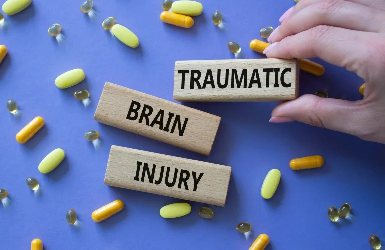 Hand holding wooden block with "TRAUMATIC" text, alongside blocks labeled "BRAIN" and "INJURY," surrounded by yellow and orange pills on blue background.