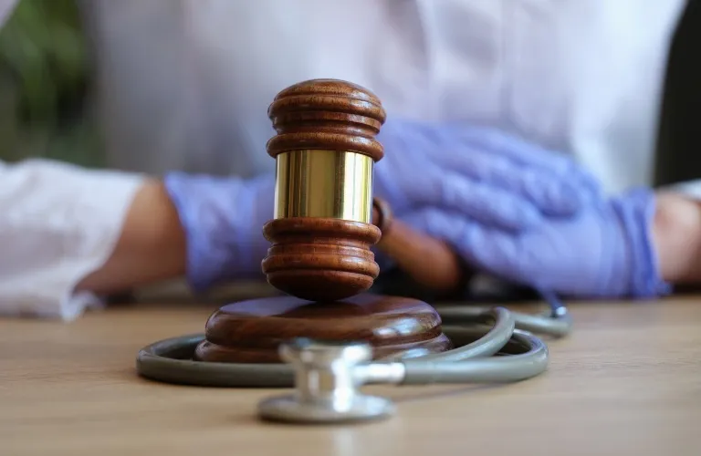 Wooden judge's gavel resting on a table with a coiled stethoscope and person wearing blue gloves in the background.