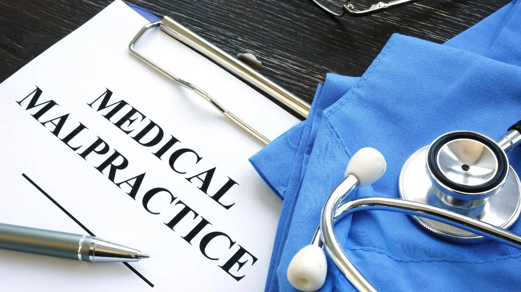 Clipboard with "Medical Malpractice" document, pen, blue medical scrubs, and stethoscope on dark wooden surface.