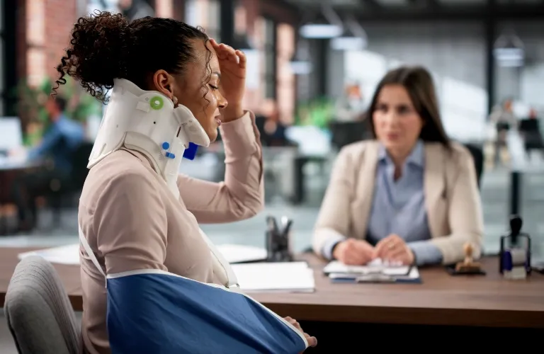 Woman with neck brace and arm sling speaking to a professional woman in an office setting.