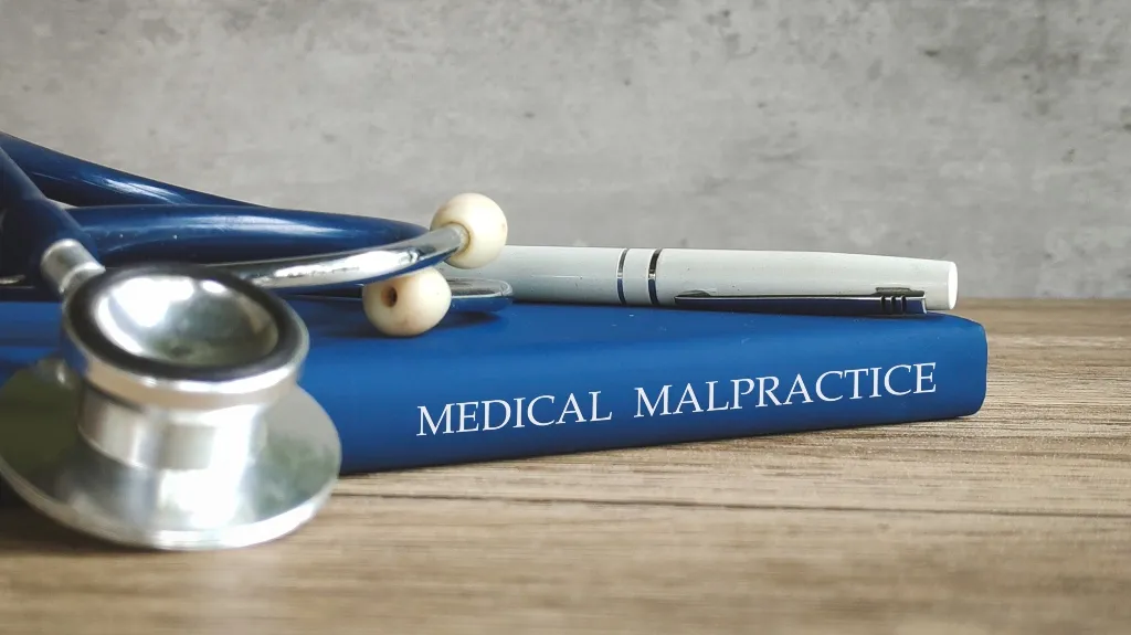 Blue book titled "Medical Malpractice" with a stethoscope and white pen on a wooden surface.
