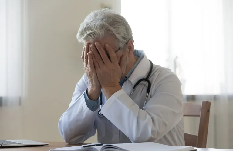 Older male doctor in white coat with stethoscope, covering face with hands, seated at desk.
