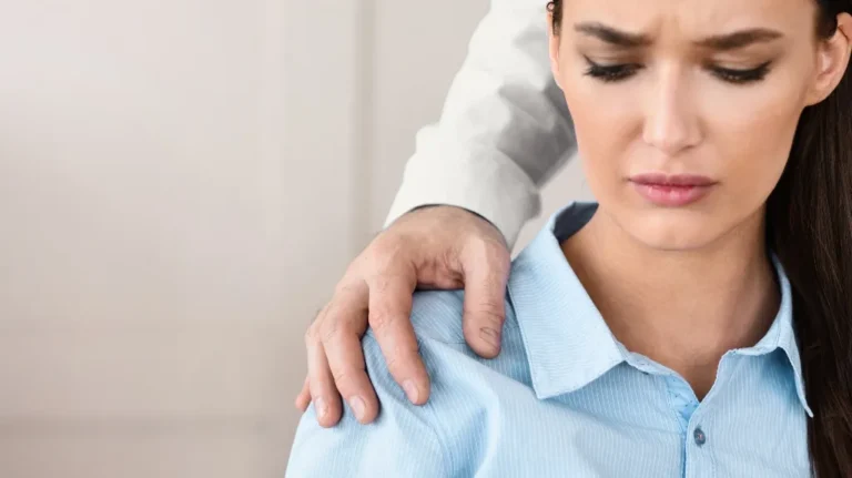 A woman in a light blue shirt looks uncomfortable as a hand rests on her shoulder.