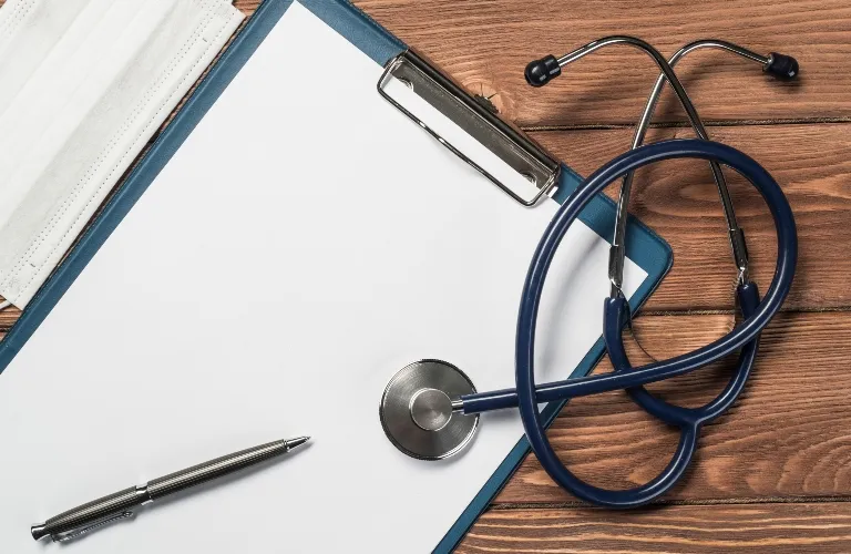 Blue stethoscope, blank clipboard with pen, and white face mask on wooden surface.