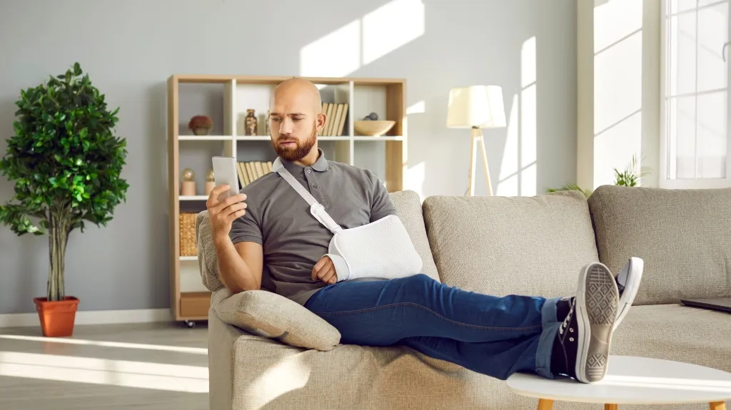 Man with arm in sling sitting on beige couch, looking at smartphone in bright living room.