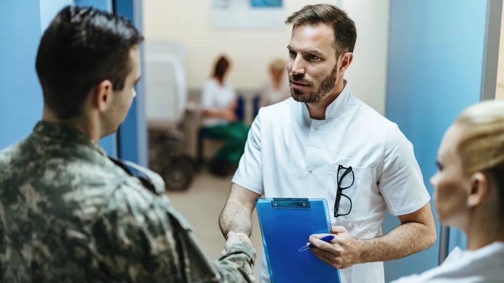 Medical professional in white coat shaking hands with soldier in camouflage uniform, holding blue clipboard.