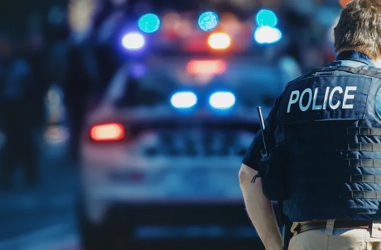 Police officer wearing a vest labeled "POLICE" standing near a patrol car with flashing red and blue lights.